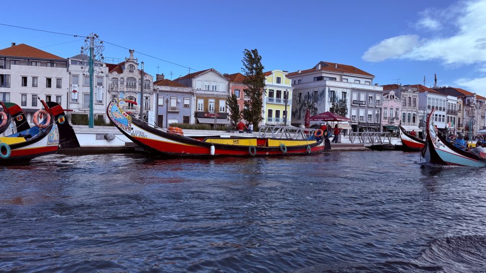 Mehrere farbenfroh bemalte, lang gezogene Boote mit hochgezogenem Bug liegen an einem Kai. Dahinter stehen helle und pastellfarbene Häuser mit Ziegeldächern, darüber ein blauer Himmel mit wenigen Wolken. Im Vordergrund kräuselt sich dunkelblaues Wasser.