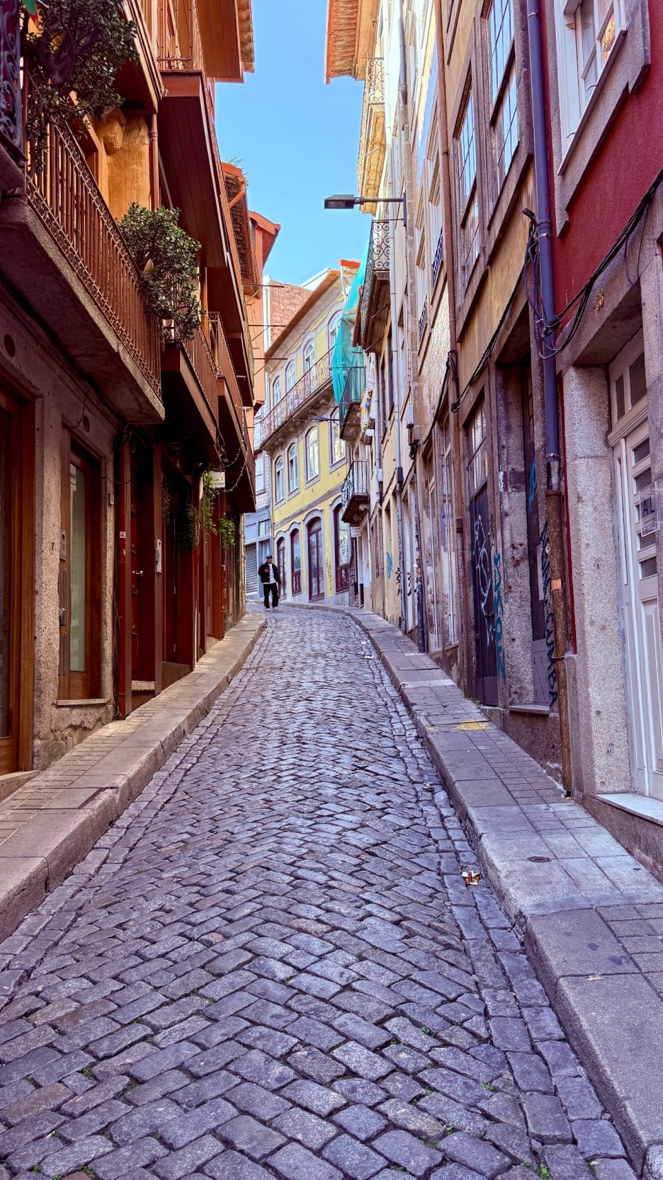 Schmale, ansteigende Gasse mit grauem Kopfsteinpflaster, links und rechts hohe Häuser in Rot, Ocker und Gelb mit Balkonen. Weit hinten geht ein einzelner Mann, darüber ein schmaler blauer Himmelsstreifen.