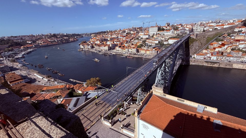 Weitwinkelaufnahme aus erhöhter Position: Eine eiserne Bogenbrücke mit zwei Ebenen führt über einen dunkelblauen Fluss. Am gegenüberliegenden Ufer drängen sich Häuser mit orangeroten Ziegeldächern den Hang hinauf. Im Vordergrund ein Dach und Mauerwerk. Blauer Himmel mit wenigen Wolken, klares Licht.