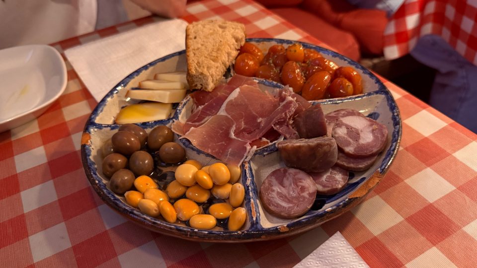 Runder Keramikteller mit blauem Rand und Fächern für Schinken, Wurstscheiben, grüne Oliven, gelbe Lupinenkerne, Käse und eingelegte orange Tomaten, dazu ein Stück Brot. Er steht auf einer rot-weiss karierten Tischdecke im warmen Licht.", "_note": ""}
Korrektur, nur das JSON:
{"text": "Kleine Platte mit Schinken, Oliven und Käse im Warmlicht. Rot-weisses Karo darunter, ein Stück Brot am Rand. Stadt erkunden macht hungrig. Und dann dieser Teller.", "alt": "Runder Keramikteller mit blauem Rand, in Fächer unterteilt: Rohschinken, Wurstscheiben, grüne Oliven, gelbe Lupinenkerne, Käse und eingeleg