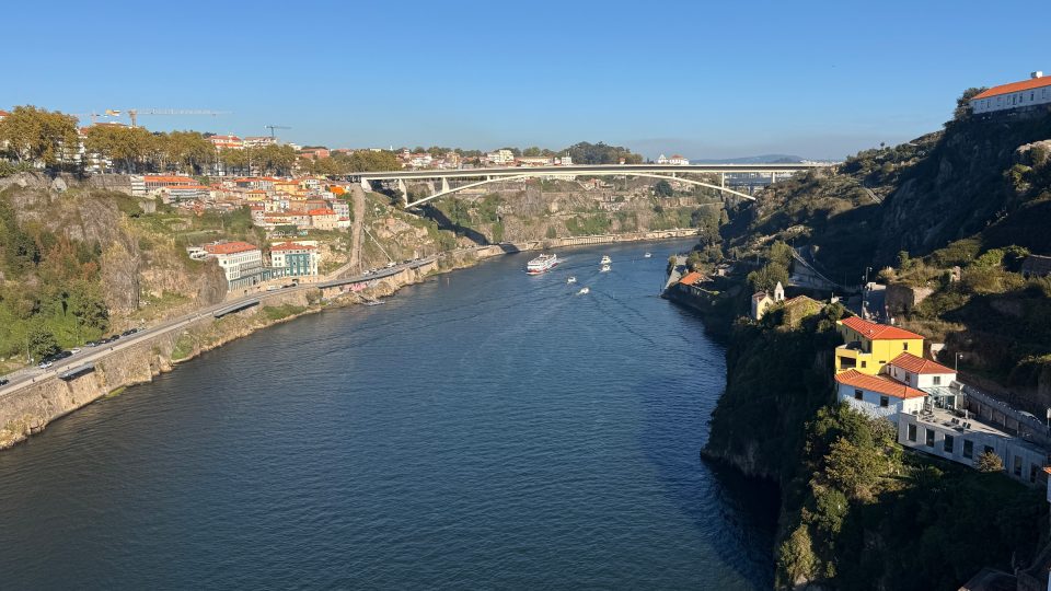 Breiter, dunkelblauer Fluss zwischen felsigen Hängen mit Häusern unter orangeroten Dächern, links eine Uferstrasse, rechts ein gelbes Haus. Im Hintergrund eine helle Bogenbrücke, auf dem Wasser einige weisse Boote, darüber wolkenloser Himmel.