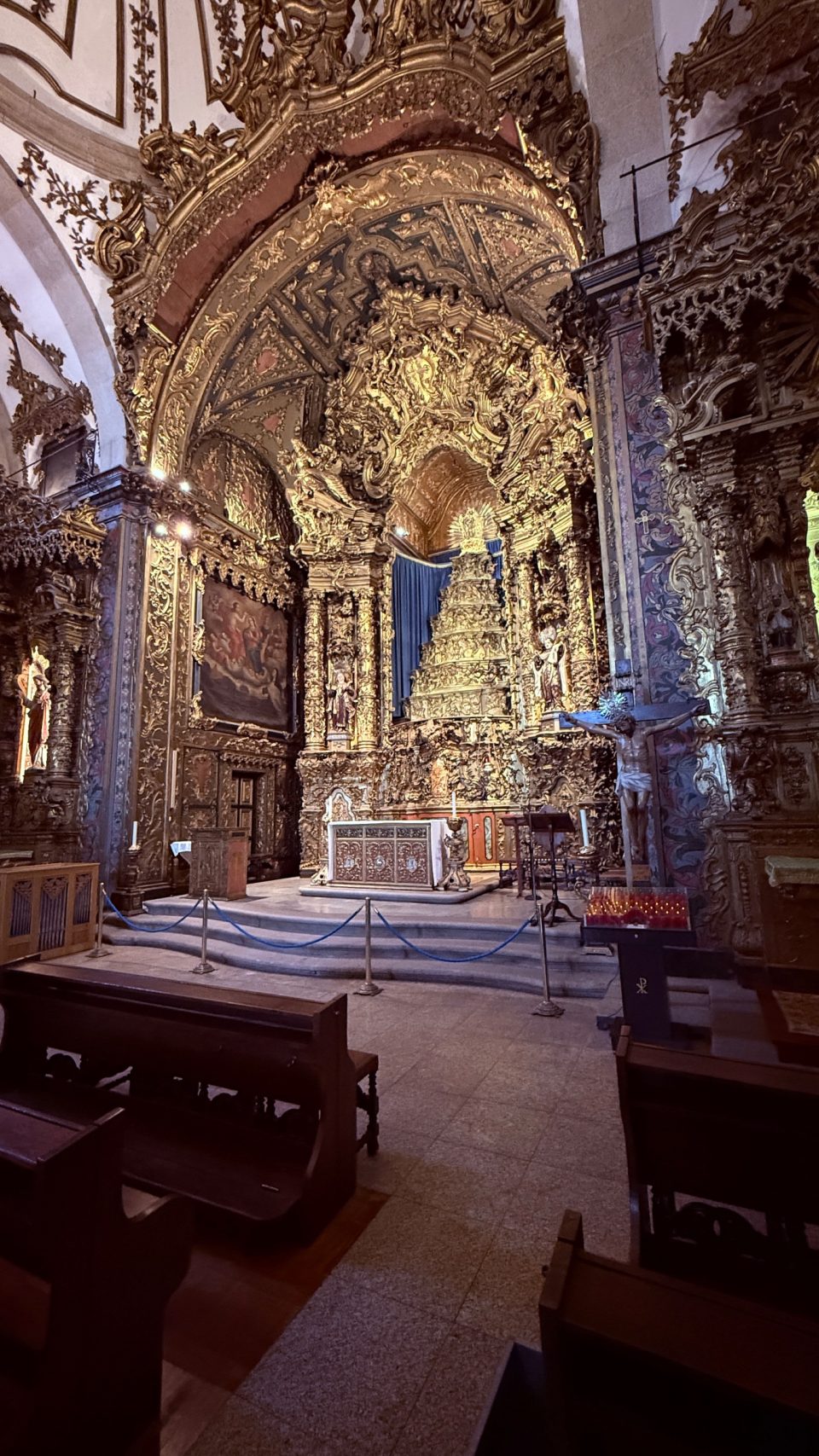 Blick in den Chor einer Kirche mit reich vergoldetem Schnitzwerk an Wänden, Bogen und Altar. Im Vordergrund stehen dunkle Holzbänke, davor ein blaues Absperrband, rechts hängt ein Kruzifix. Das Licht ist warm und gedämpft.